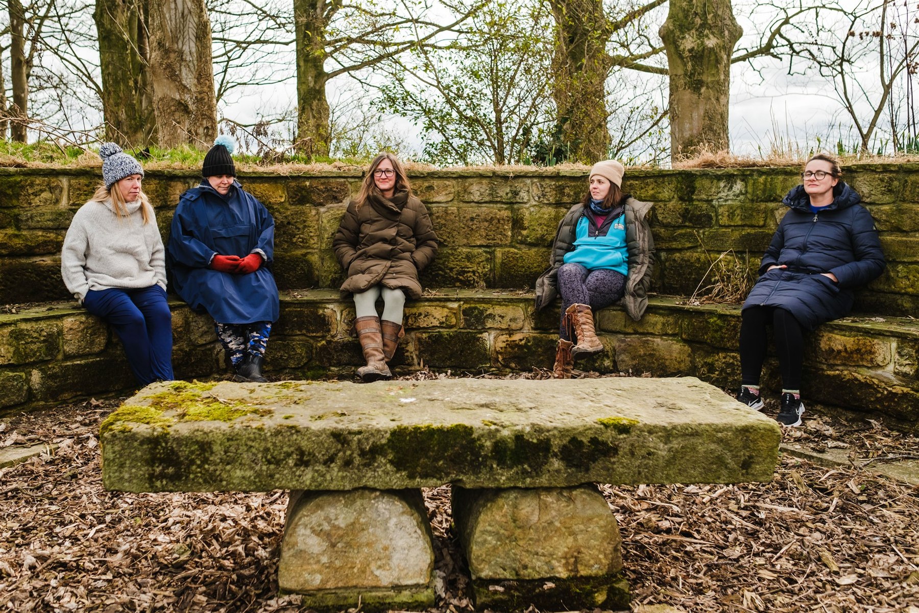 Five people taking a quiet moment on curved stone seating beneath the trees at Patty’s Barn